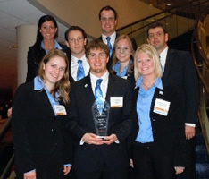 Eight students dress in business outfits with a blue theme. The front student is holding a glass plaque