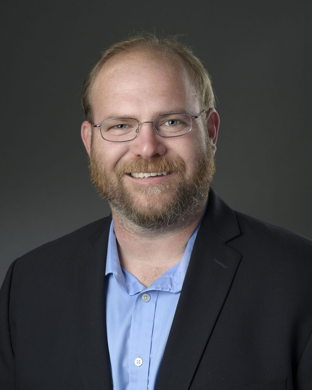 White male with glasses in a blue shirt and black sports jacket.