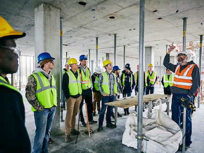 Photo of class of studente touring construction site, with professor pointing to concrete ceiling.