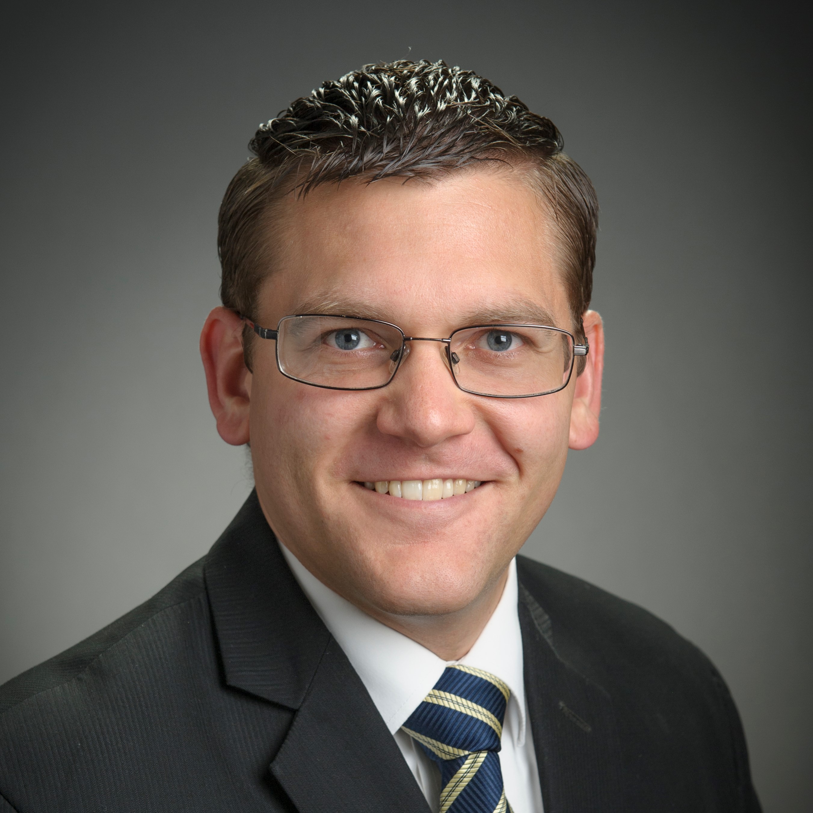 Caucasian man with short dark hair wearing a suit, tie and glasses with a gray background.