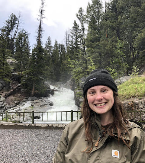 Samantha Milner stands in front of steep whitewater rapids and evergreen trees
