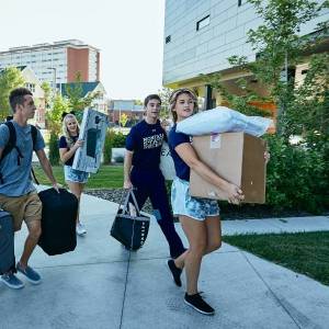 four students carrying bedding and boxes into a building