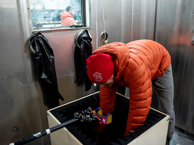 MSU student in puffy orange coat bending over to handle experiment in dark box inside freezer chamber