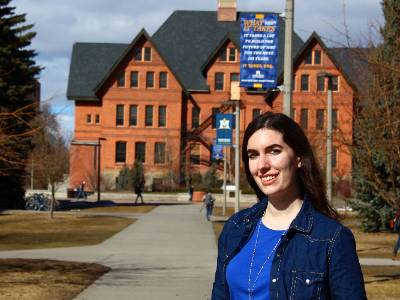 Kailyn is in a blue shirt and jean jacket in front Montana Hall with MSU and What It Takes banner overhead.