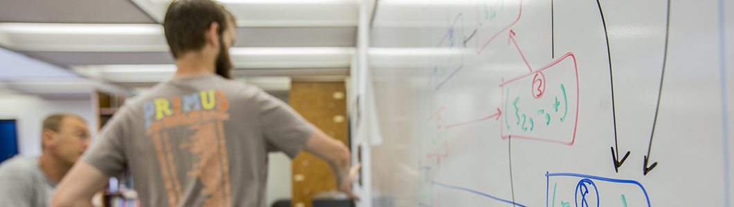 A young man and his mentor discuss calculations on a white board.