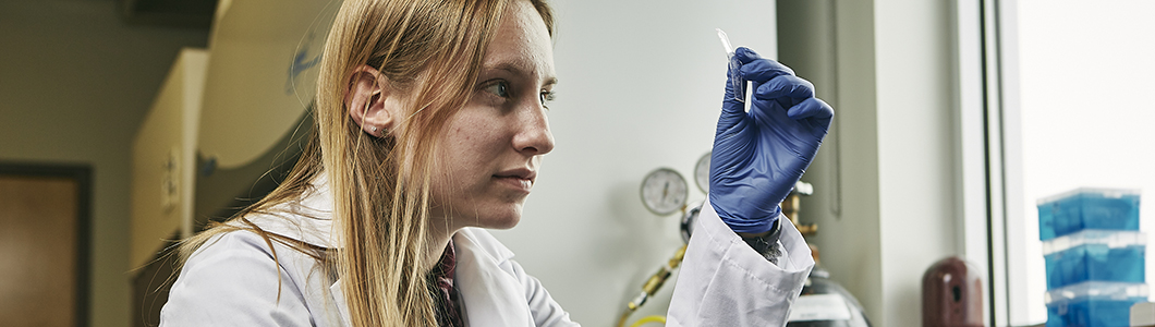 A young woman in a lab coat and rubber gloves observes a sample in a test tube.
