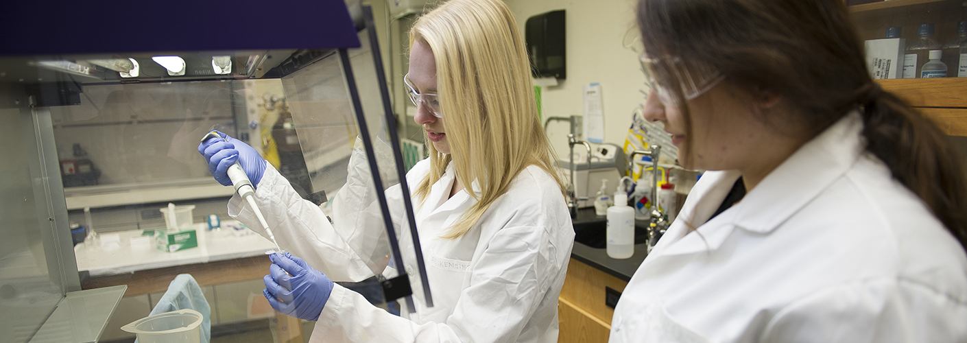 Two young women are in a lab; one, with blond hair, is pipetteing while the other looks on.