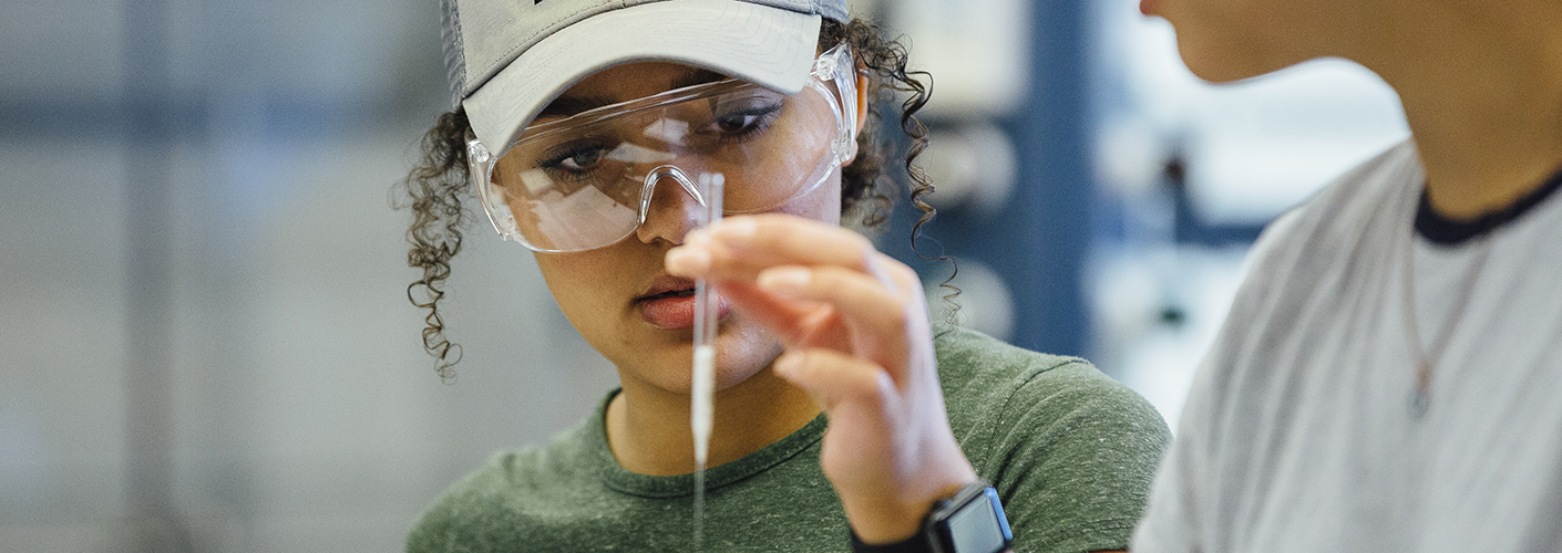 A young African American woman with curly hair in a ball cap inspects a pipette.