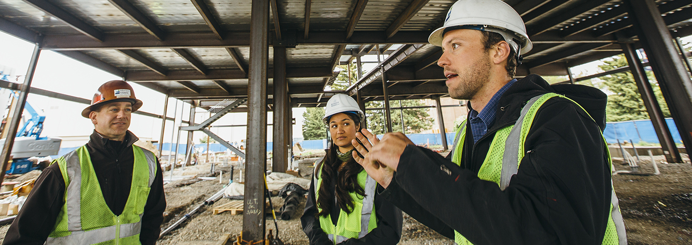 Three figures (two men and one woman) in hard hats talk in the midst of a construction site with iron beams surrounding them.