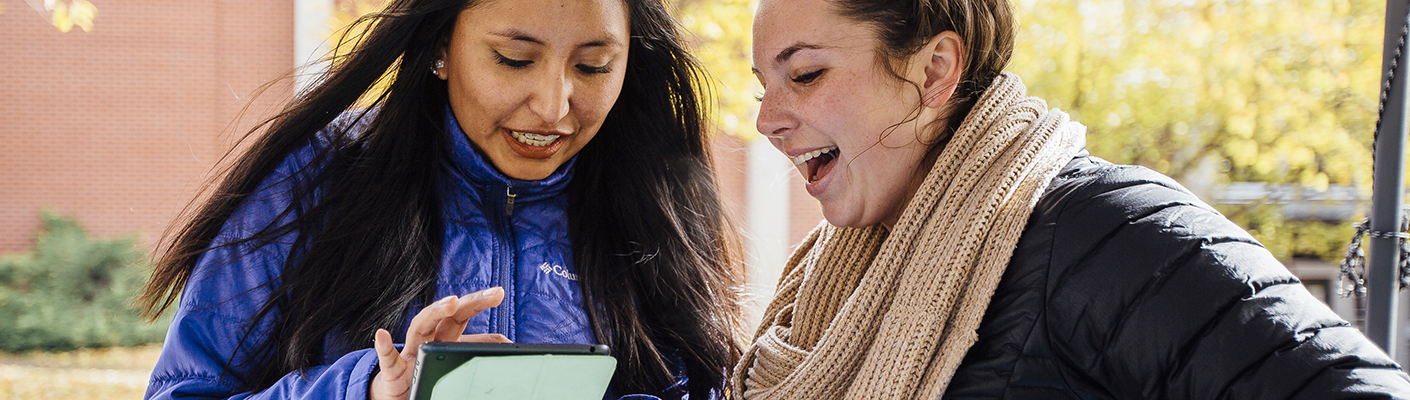 Two female students hold a discussion over a small tablet outside.