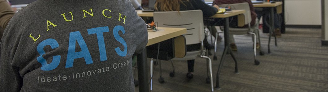 A student sits in a classroom, their "Launch CATS" t-shirt displayed to the viewer.