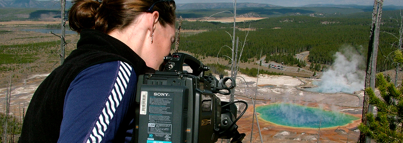 A young woman is bent over a camera, pointing towards the Great Prismatic Spring (a pool of water that is deep cerulean, then ringed with green, and finally fading towards orange) in Yellowstone National Park.