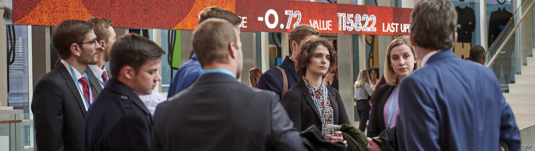 A group of students in business formal attire stand in the lobby of Bloomsburg Media prior to a tour of the facility, a stylized stock display in the background.