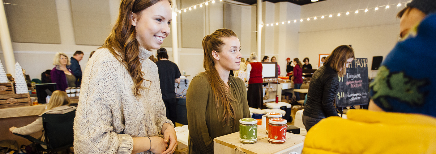 Two young women address customers that are looking at their product: fermented food in jars.