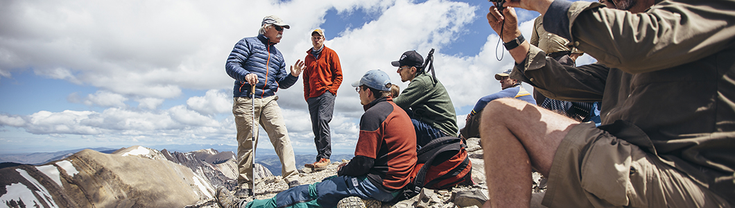 A group of students surround a professor as he lectures at the summit of a mountaintop.