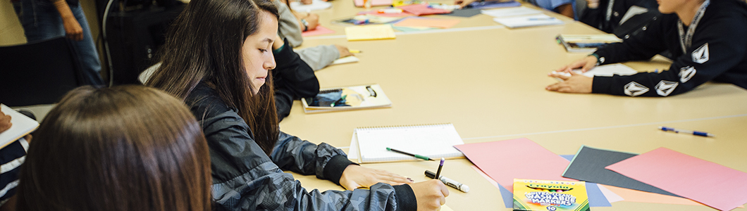 A young woman sits at a table with elementary school students, writing on a piece of paper.