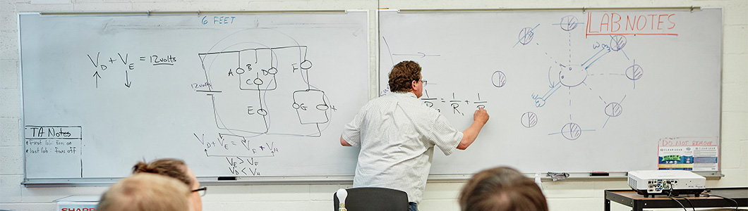 A teacher gestures at the chalkboard while students look on.