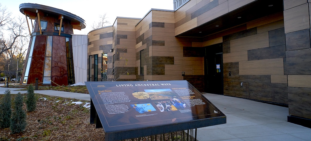 The American Indian Hall is shown from its side with an interpretive sign in the foreground that reads "Living Ancestral Ways."