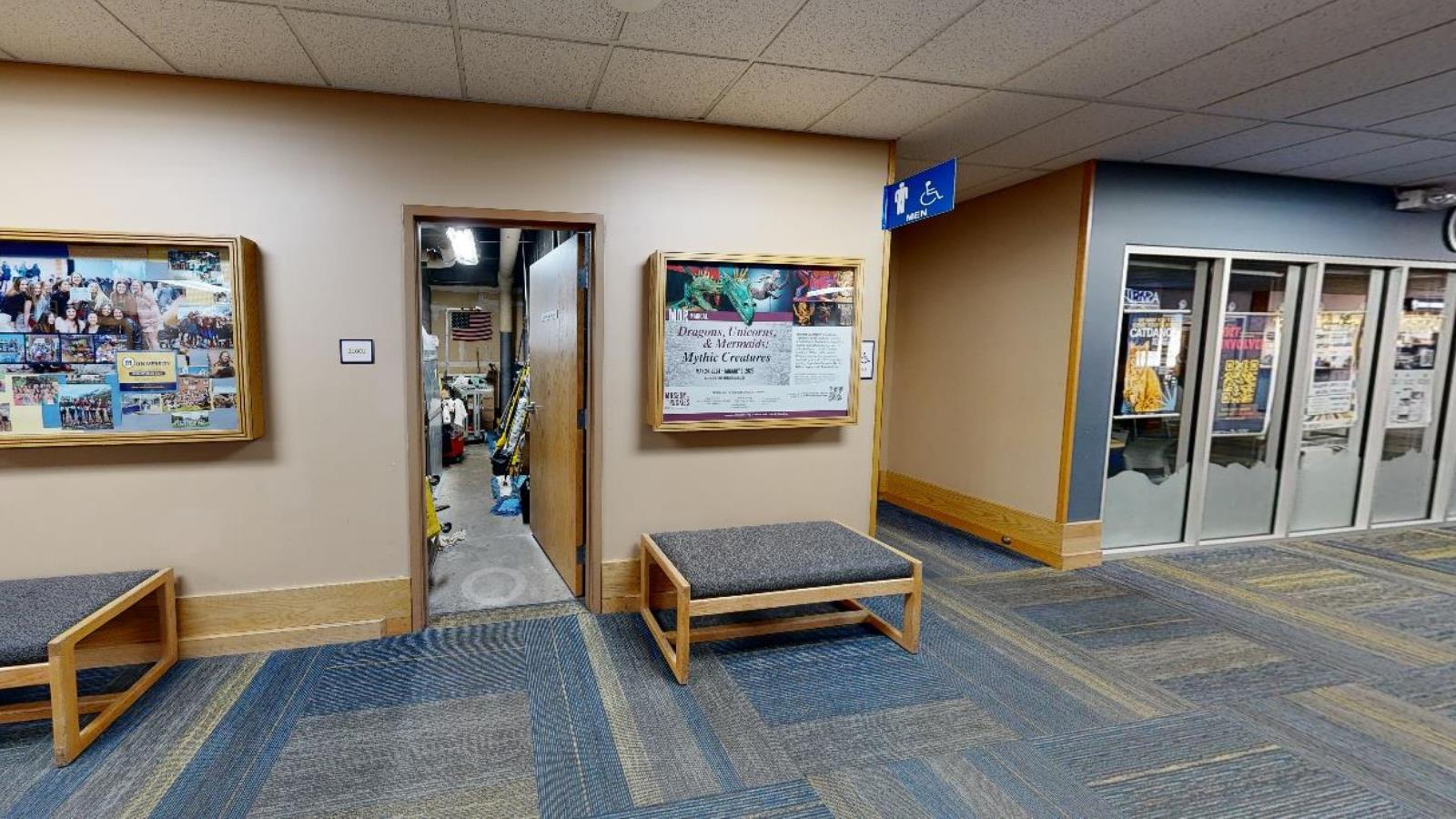 A hallway in the student union building with beige walls and blue carpet. A framed collage of student photos is mounted on the left wall, and an open storage room is visible with equipment inside. A blue accessibility sign marks a men’s restroom nearby.