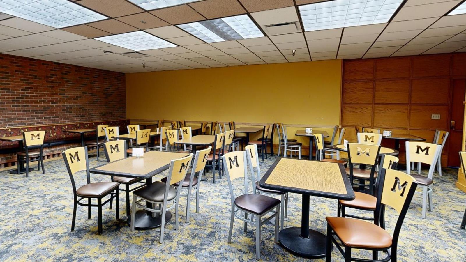 A dining area with yellow walls, patterned carpet, and wooden furniture. Chairs are branded with an "M" logo, and seating options include tables with individual chairs and a cushioned bench along the wall. The ceiling consists of drop tiles with fluorescent lighting.