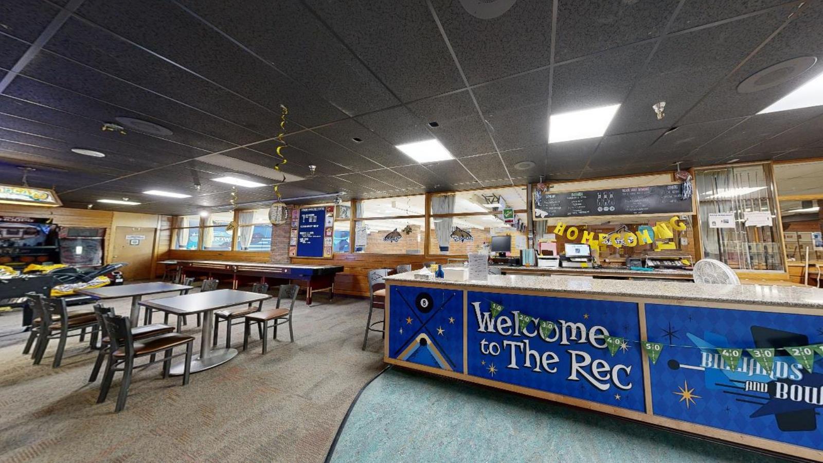 A student recreation center featuring a check-in counter with "Welcome to The Rec" signage. The space has pool tables, arcade games, and a seating area with tables and chairs. Decorations include Homecoming-themed banners and blue and gold accents.