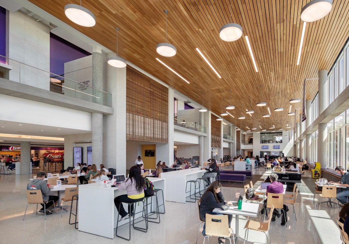 A spacious, high-ceilinged university student center with large windows allowing natural light to fill the space. Students are seated at communal tables, high-top counters, and small group tables, engaging in studying, eating, and socializing. The wooden ceiling, concrete columns, and glass-paneled balconies give the space a contemporary design, and a food court is visible in the background.