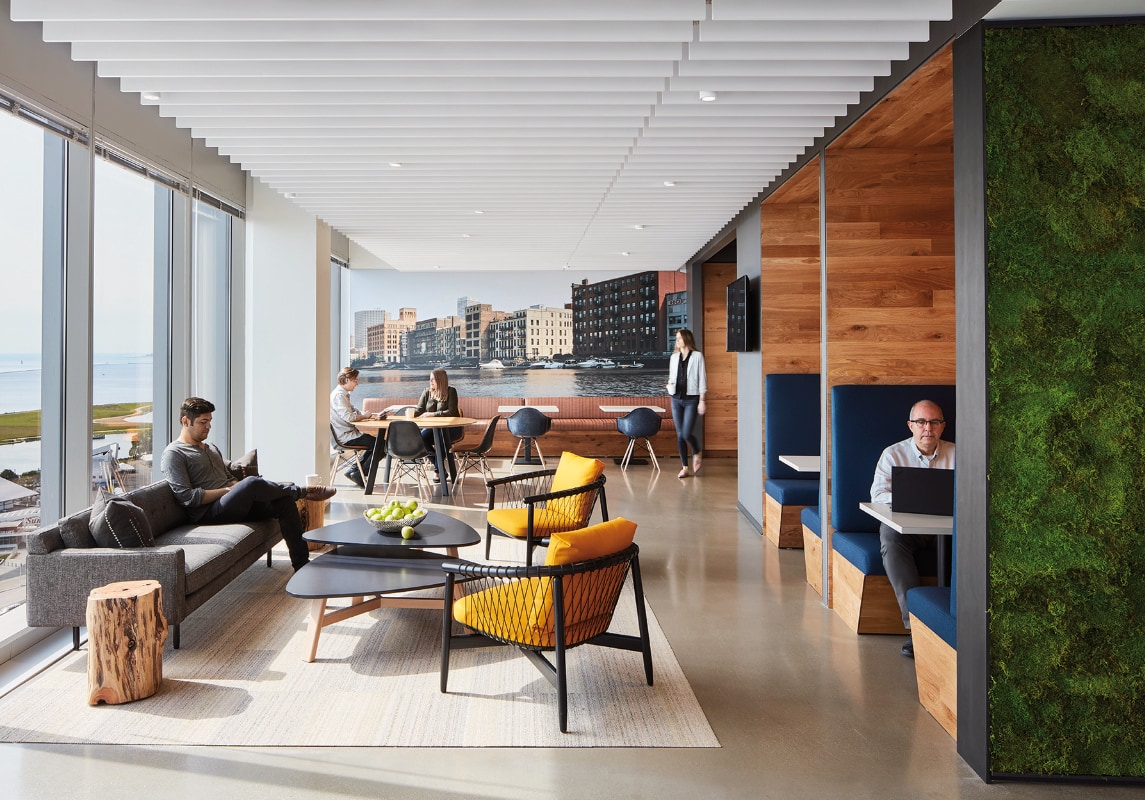 A modern, open-concept office space with large windows offering a view of a waterfront landscape. The room has various seating options, including sofas, armchairs with yellow cushions, and private booth seating with blue upholstery. A moss-covered wall adds a natural touch, while several people are seen working on laptops or conversing at tables.