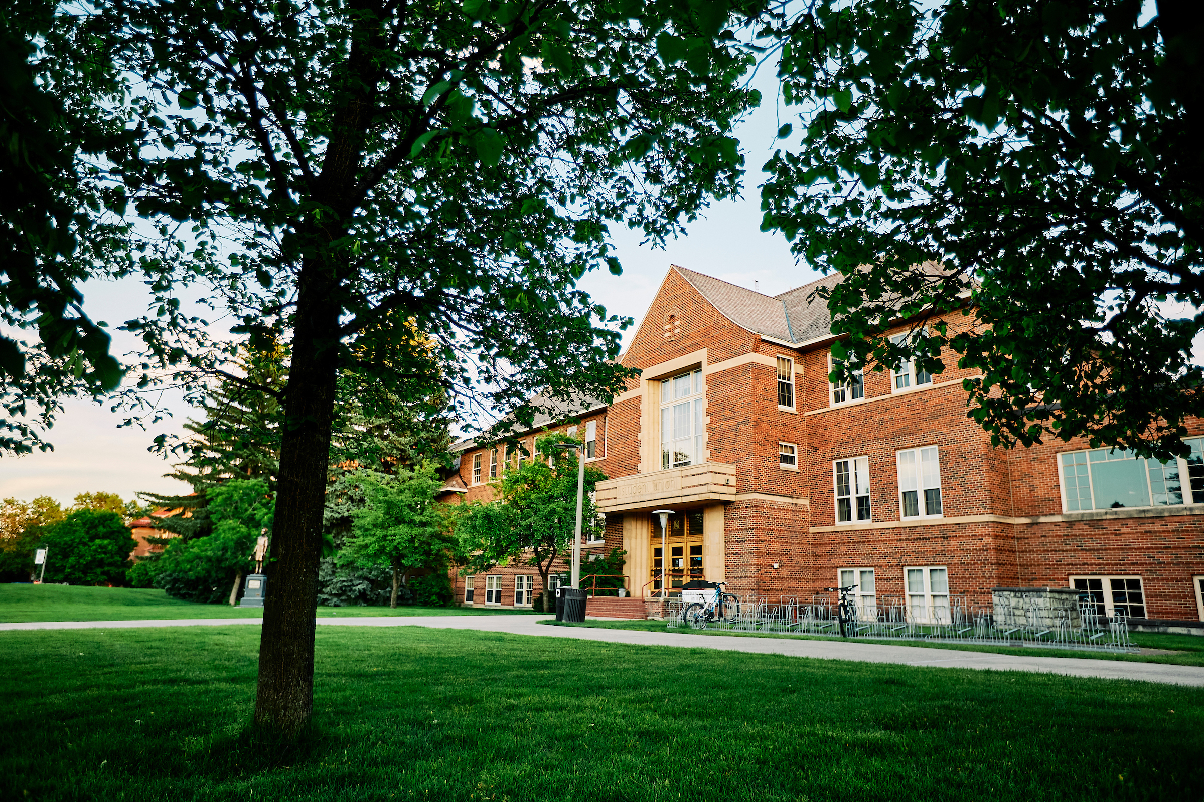 A historic red brick university building with large windows and a central entrance framed by beige stone columns. The building is surrounded by lush green grass, mature trees, and a paved walkway. Bicycles are parked in racks near the entrance, and a statue is visible in the background. The setting appears to be late afternoon or early evening, with soft natural lighting filtering through the trees.