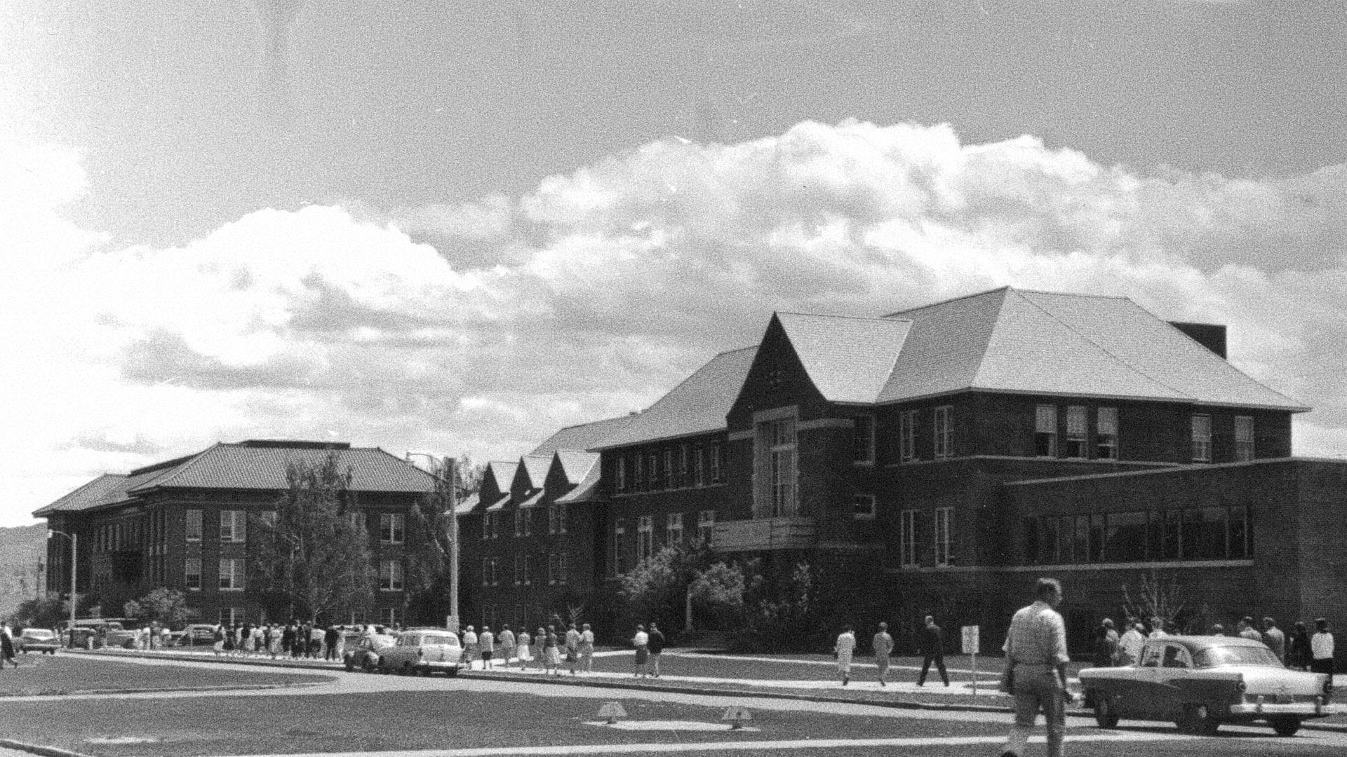 A black-and-white historical photograph captures a scene on a college campus during daytime. The image features large brick buildings with pitched roofs and rows of windows, likely mid-20th century academic or administrative structures. A group of people, dressed in mid-century attire, walk along the sidewalks and cross the street, engaging in daily campus activities. Classic automobiles from the era, including a prominent vintage car in the foreground, are parked along the road. The background showcases a partly cloudy sky, adding depth to the scene.