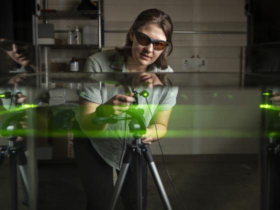 A woman, wearing laser protective glasses and a mint green quilted shirt, leans over a green laser that is pointing into a tank of water. The beam of the laser illuminated a level plain of the water and the glass sides of the tank reflect portions of the  | MSU photo by Colter Peterson