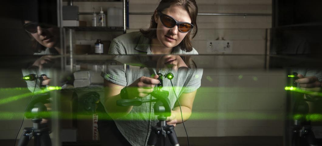 A woman, wearing laser protective glasses and a mint green quilted shirt, leans over a green laser that is pointing into a tank of water. The beam of the laser illuminated a level plain of the water and the glass sides of the tank reflect portions of the 