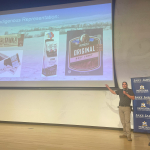 A man standing near a lecturn presents a powerpoint presentation about his business.