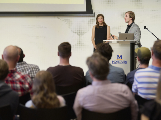 Two presenters stand behind a podium in front of an audience.  | Colter Peterson