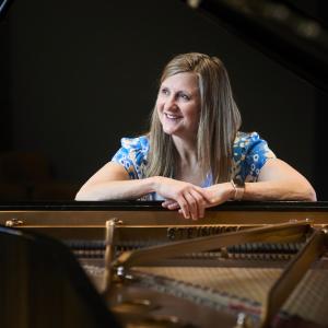 A woman wearing a blue dress leans on the opening of a grand piano. The stings of the piano are seen in the foreground
