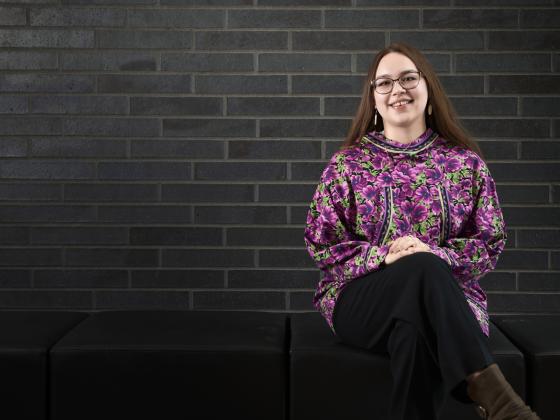 A woman wearing a pink flowery patterned shirt sits with her legs crossed and hands on her knees while smiling at the camera. She is sitting on a soft, black bench in front of a dark grey brick wall. | MSU photo by Colter Peterson