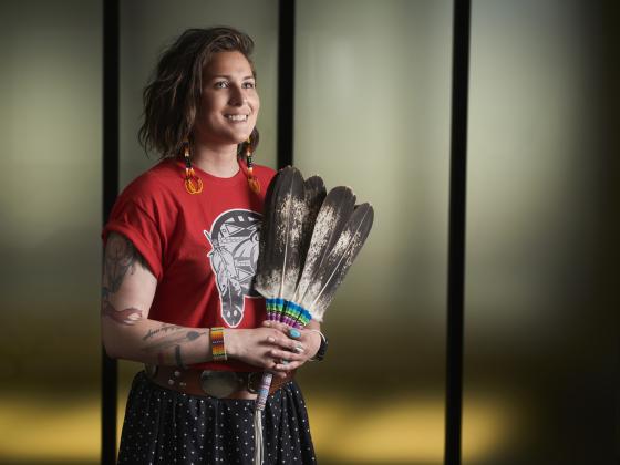 A brown haired woman wearing a red t-shirt with the American Indian Council logo on it and a black skirt with polka-dots smiles while looking off to the top right of the frame, she is holding a eagle feather fan with both hands. The background is an out o | MSU photo by Colter Peterson