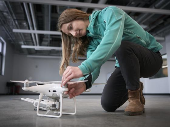 A woman wearing a thin blue jacket and black pants kneels on a concrete floor while attaching a prop to a white plastic drone.