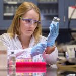 Lisa Keller works in a lab setting with glass vials while wearing a lab coat and other PPE