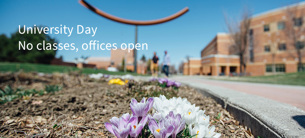 Flowers poke through the dirt in a planter bed next to a sidewalk. A blue sky overlook at a college campus in the background. | MSU