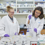 Two women inside a chemistry lab, wearing lab coats, safety glasses and vinyl gloves, work with pipets on a lab bench.