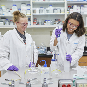Two women inside a chemistry lab, wearing lab coats, safety glasses and vinyl gloves, work with pipets on a lab bench.