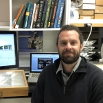 A man sits in front of a computer screen and an insect display box