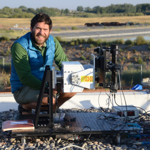 Man smiling in blue vest crouching outside by scientific equipment.