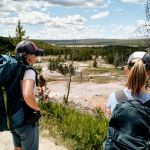 Two female scientists wearing backpacks gaze out upon Yellowstone hot spring amid pines.