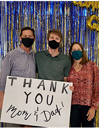 Parents with their student hold a sign that says THANK YOU Mom and Dad! They are standing in front of shiny blue and gold streamers