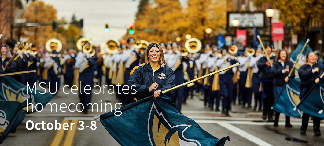 A cheerleader twirling a flag marches down a street in front of a marching band during an autumn homecoming parade.  | MSU