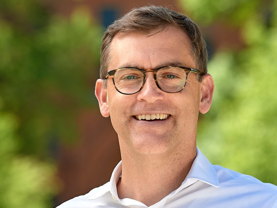 Brock Tessman, wearing glasses and a white button-up shirt, smiles outdoors in front of blurred green foliage and a brick building. Overlaid text on the left reads: “MSU welcomes President Brock Tessman.” | MSU
