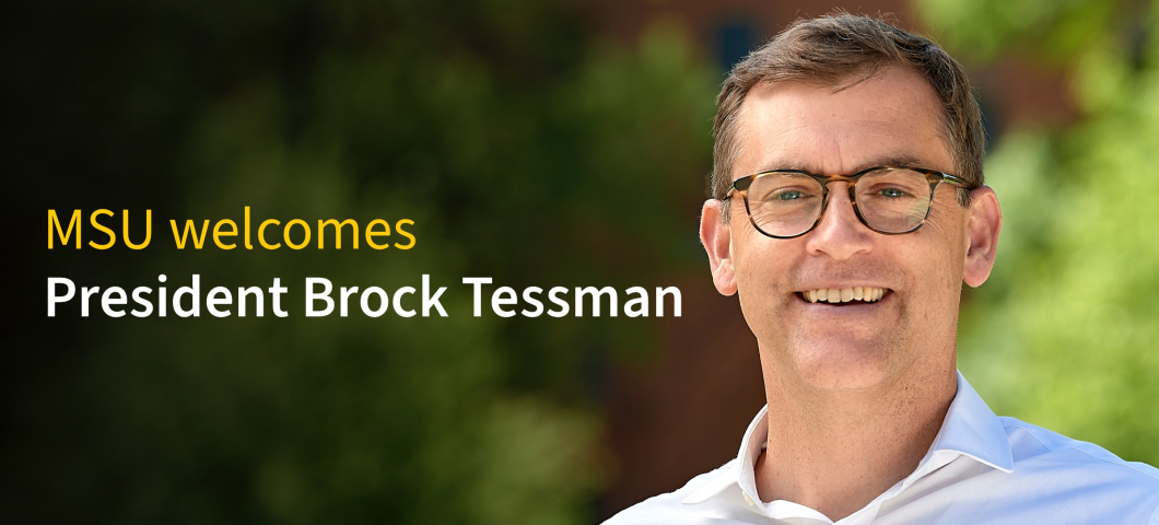 Brock Tessman, wearing glasses and a white button-up shirt, smiles outdoors in front of blurred green foliage and a brick building. Overlaid text on the left reads: “MSU welcomes President Brock Tessman.” | MSU