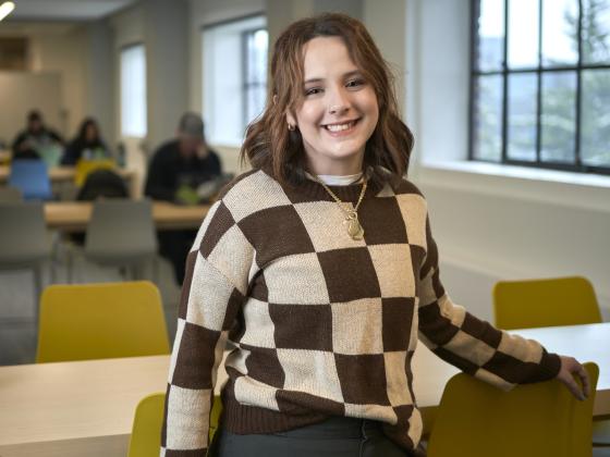 A young woman wearing a brown and tan checkered sweater and a gold necklace and pendant smiles at the camera while leaning back against a table. Students are studying in the background, out of focus. | MSU photo by Colter Peterson