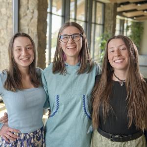 A trio of women stand together arm-in-arm while looking at the camera